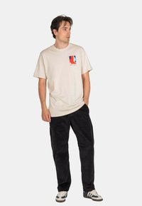 Mann trägt ein beigefarbenes T-Shirt mit Grafik, schwarze Hose und weiße Sneaker. Er steht mit einer Hand in der Tasche und blickt seitlich vor einem einfarbigen Hintergrund.