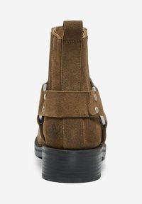 Bottes de cheville en suède marron avec une sangle et des accents métalliques, dotées d'un talon noir uni et d'une surface texturée. Présentées de la vue arrière.