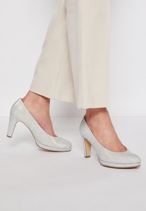 Ben i lyse beige bukser iført sølvglitrende højhælede sko med lukket tå, der står på en hvid overflade.