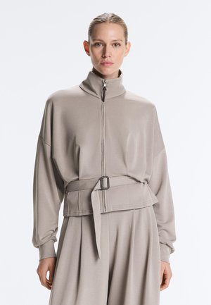 Žena oblečená v svetlo taupe top s golierom na zips a široké nohavice s opaskom v rovnakom odtieni, stojaca pred jednoduchým svetlým pozadím.