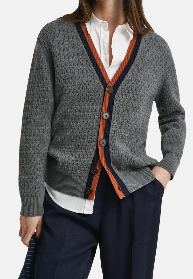 Femme portant un cardigan gris texturé avec des bordures bleu marine et orange, par-dessus une chemise blanche boutonnée et un pantalon bleu marine, la main dans la poche.