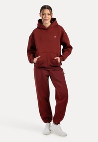 Felpa con cappuccio bordeaux con tasca frontale, abbinata a pantaloni sportivi tapered coordinati. La trama del tessuto appare morbida, stile casual. Sneakers bianche.