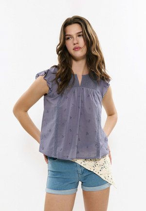 Jeune femme aux longs cheveux ondulés portant un chemisier violet sans manches et un short en jean bleu avec un détail au crochet blanc.