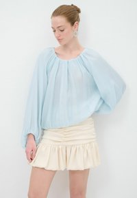 Helle blaue transparente Bluse mit Rundhalsausschnitt und langen Ballonärmeln, kombiniert mit einem cremefarbenen Minirock mit Rüschen und gerafften Details.