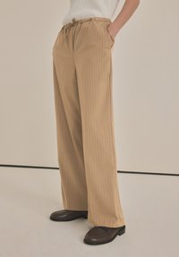 Pantaloni beige a righe con elastico in vita e coulisse, dotati di tasche laterali e vestibilità rilassata, abbinati a scarpe marrone scuro.