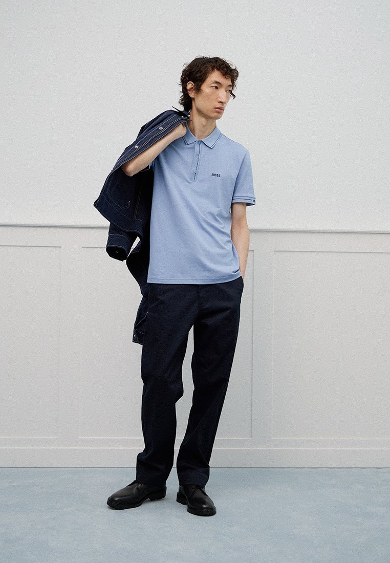 Junger Mann in hellblauem Poloshirt und dunkler Hose, der eine dunkle Jacke über die Schulter hält, steht vor einer hellen Wand.