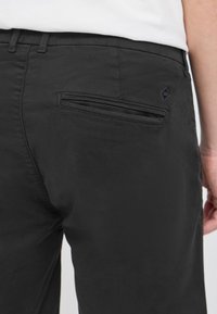 Gros plan d'une personne portant un pantalon noir avec une poche passepoilée à l'arrière et une chemise blanche rentrée.