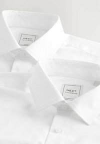 Chemises blanches avec col à boutons, présentant une texture lisse et une étiquette indiquant "NEXT TAILORING." Deux chemises superposées.