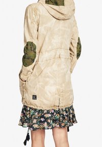 Beżowa kurtka z kapturem, z zielonymi łatami w kamuflażu na łokciach oraz ściągaczem w talii. Na dole widoczna kwiecista spódnica o warstwowej konstrukcji.