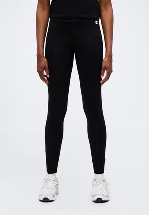 Benen met zwarte strakke leggings met het Champion-logo op de linkerheup, witte sokken en witte sportschoenen tegen een effen lichte achtergrond.