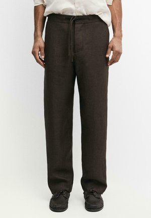 Uomo che indossa pantaloni marrone scuro a gamba dritta con coulisse in vita e scarpe nere, in piedi davanti a uno sfondo bianco semplice.