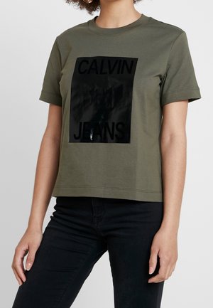 Person trägt ein olivgrünes kurzärmeliges T-Shirt mit einem glänzenden schwarzen Rechteck, das den Text "Calvin Jeans," zeigt, kombiniert mit einer schwarzen Hose.