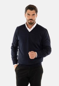 Maglione blu navy a V in un tessuto testurizzato, abbinato a una camicia bianca con colletto, indossato con pantaloni neri. Presenta polsini e orlo a coste.