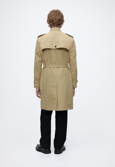 Personne aux cheveux bouclés portant un trench-coat beige, un pantalon noir et des chaussures noires, debout de dos sur un fond blanc uni.