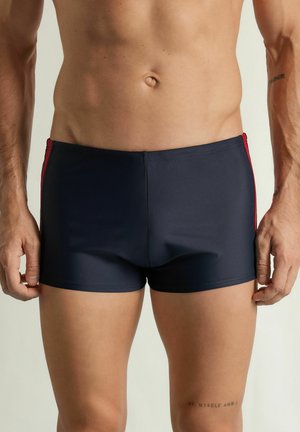 Męski tors w granatowych kąpielówkach z czerwonymi paskami po bokach, stojący z rozluźnionymi rękami na jednolitym jasnym tle.