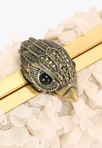 Pochette beige con tessuto floreale strutturato, hardware dorato e chiusura decorativa a forma di uccello con accenti neri e iridescenti.