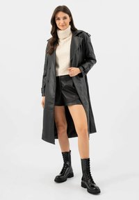 Cappotto lungo in pelle nera con design doppiopetto, ampie revers e grandi tasche. Indossato sopra un maglione in maglia color crema e pantaloni corti neri.