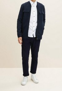 Chaqueta bomber de navy con cremallera y dos bolsillos en el pecho, camisa blanca de botones y pantalones oscuros; acompañada de zapatillas blancas. Tejido de textura suave.
