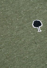 Tela verde oliva con apariencia texturizada, que presenta un pequeño diseño de árbol bordado en negro contorneado en blanco.