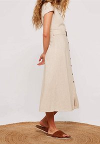 Robe en lin beige clair avec des manches courtes, des boutons à l'avant et une taille cintrée. Elle présente une jupe évasée et une fente sur le côté. Portée avec des sandales tressées marron.