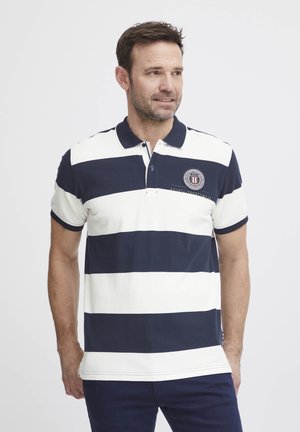 Polo en rayas azul marino y blancas, de mangas cortas y con cuello. Presenta un logo bordado en el lado izquierdo del pecho y acentos oscuros contrastantes.