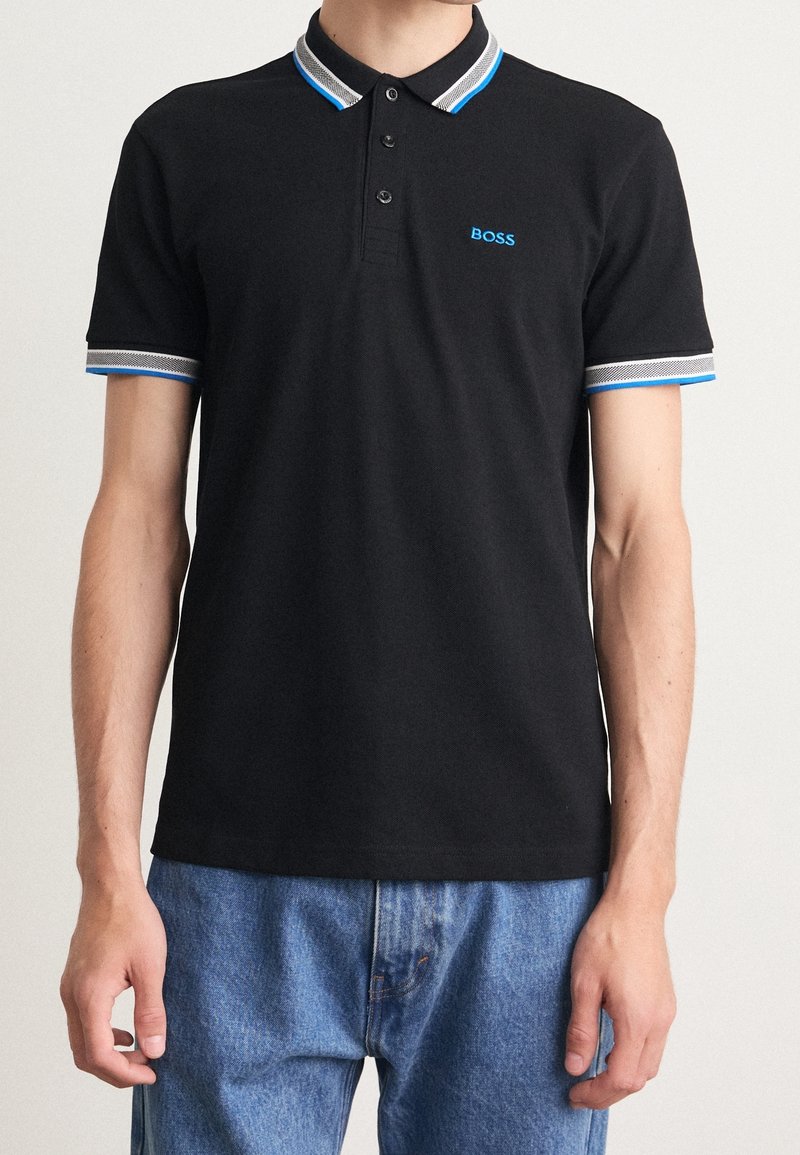 Homme portant un polo noir avec des bordures bleues et blanches au niveau du col et des manches, logo "BOSS" brodé en bleu sur la poitrine, associé à un jean bleu.