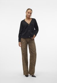 Schwarze Strickjacke mit V-Ausschnitt, zwei vorderen Taschen und Knopfverschluss, kombiniert mit weiten Hosen im Leopardenmuster und schwarzen Absatzschuhen.