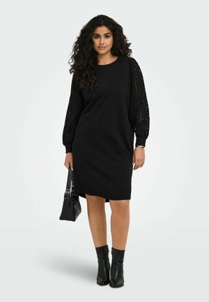 Femme aux cheveux bouclés portant une robe noire longueur genou avec des manches en dentelle, tenant un sac à main noir et portant des bottines noires, debout avec assurance.