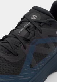 Shoe sport negru cu partea superioară din plasă, accente texturate și branding gri. Prezintă vârf rotunjit, sistem de închidere cu șireturi și elemente de design albastre subtile.