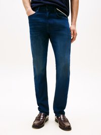 Jeans de denim azul escuro com um corte slim, apresentando um design de cinco bolsos e um desbotado subtil. Usados com mocassins castanhos escuros.