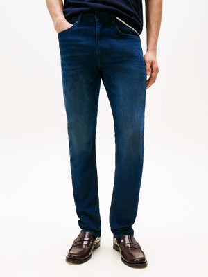 Jeans de denim azul escuro com um corte slim, apresentando um design de cinco bolsos e um desbotado subtil. Usados com mocassins castanhos escuros.