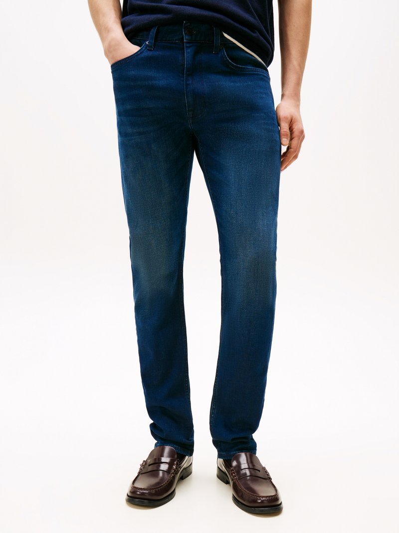 Jeans de denim azul escuro com um corte slim, apresentando um design de cinco bolsos e um desbotado subtil. Usados com mocassins castanhos escuros.
