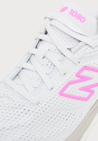 Κοντινό ενός λευκού αθλητικού παπουτσιού New Balance 1080 με ροζ λογότυπο και σήμανση, με διχτυωτό ύφασμα και λευκά κορδόνια διασταυρωμένα.