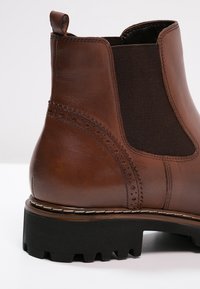 Bottine en cuir marron avec panneaux élastiques sur les côtés, détails de brogue et semelle en caoutchouc noir texturé. Équipée d'une boucle pour un enfilage facile.