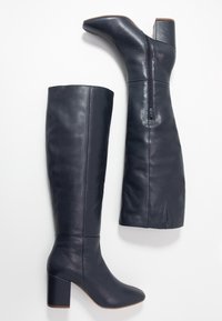 Bottes hautes en cuir bleu foncé, avec un bout pointu, un talon bloc et un détail de fermeture éclair. Caractérisées par un design élégant et des fentes latérales en haut.