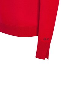 Maglione rosso lavorato a maglia con orlo e polsini a costine. Presenta spacchi laterali sul fondo e un piccolo logo nero sul polsino. Texture morbida.