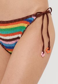 Bas de bikini rayé en matériau crochet dans des couleurs vives. Présente des liens bruns ajustables avec des accents colorés aux extrémités.