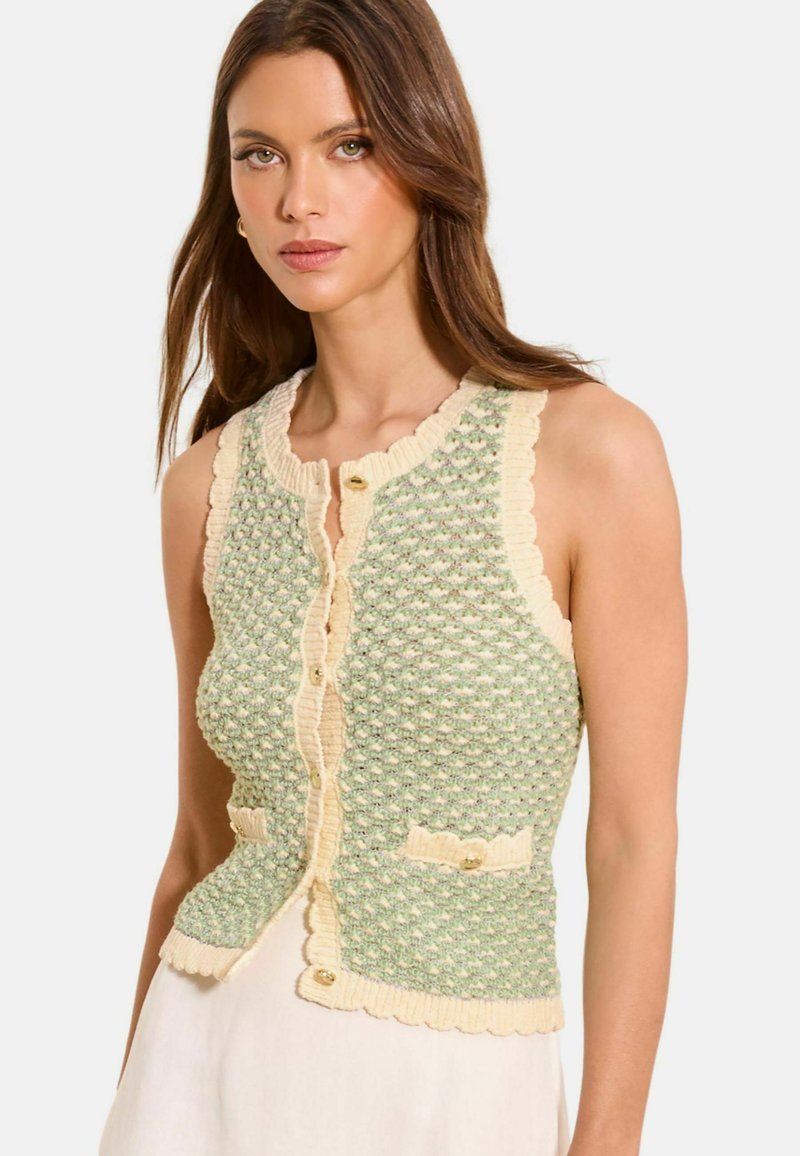 Femeie purtând un vestă tricotată fără mâneci, texturată, în verde și crem, cu margini cu volane și nasturi aurii, asortată cu o fustă albă.