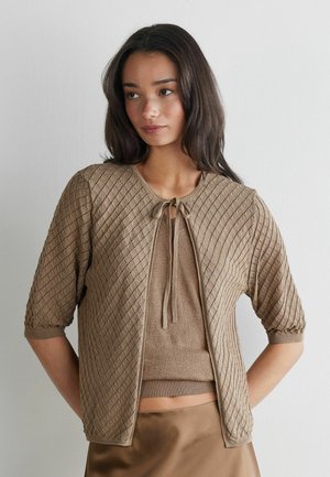 Junge Frau mit langen dunklen Haaren, die eine beige gestrickte Strickjacke über einem passenden ärmellosen Oberteil und einem braunen Rock trägt, steht vor einer weißen Wand.