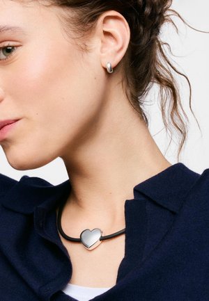 Femme portant un pendentif argenté en forme de cœur sur un collier en cordon noir et des boucles d'oreilles créoles argentées assorties, vêtue d'un haut bleu marine.