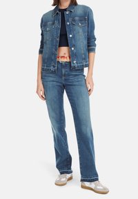 Femme portant une veste en jean bleue et un jean, un top court noir dévoilant le ventre, une ceinture beige et des baskets blanches avec des accents beiges.