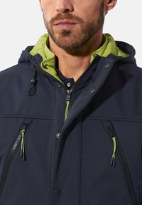 Navy jacket with a green hood, featuring zippers and drawstrings. Smooth fabric, with side pockets and contrasting zipper details.