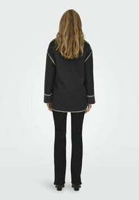 Cappotto di lana nero con maniche lunghe, caratterizzato da una cucitura decorativa bianca lungo le cuciture e un design in stile tunica. Abbinato a pantaloni neri aderenti.