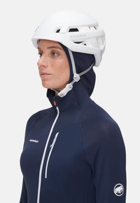Biały kask wspinaczkowy z otworami wentylacyjnymi, połączony z granatową kurtką zapinaną na zamek z kieszenią na piersi oraz logo na rękawie.