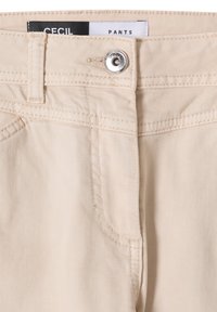 Pantalon beige avec poches cousues, passants pour ceinture et fermeture par bouton argenté marqué "CECIL", avec une étiquette de marque visible à l'intérieur de la ceinture.