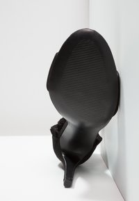 Chaussure noire à talon haut positionnée semelle vers le haut contre une surface d'angle blanche, montrant une semelle en caoutchouc texturée et une fine pointe de talon.