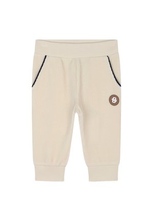 Jogging en velours côtelé beige clair avec des coutures bleu marine, deux poches latérales et un petit patch logo marron à l'avant. Texture douce, taille élastique.