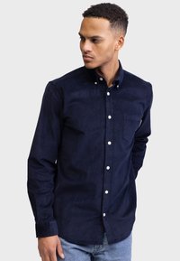 Camisa de pana en color navy con cuello abotonado, bolsillo frontal y botones blancos. Presenta una tela texturizada y mangas largas. El modelo lleva unos jeans claros.