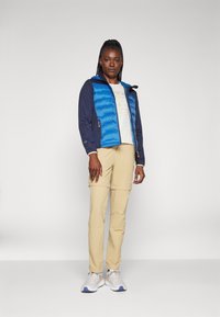 Casaco puffer azul e marinho com fecho de correr, calças cargo bege, t-shirt branca com um grafo circular e sapatilhas cinzas.