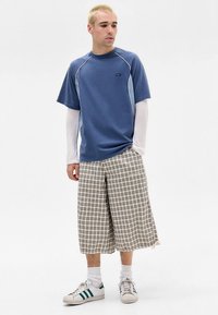 Kék, bő fazonú póló kontrasztos világoskék varrásokkal, fehér hosszú ujjú aláöltözővel és bézs kockás rövidnadrággal. Fehér sportcipő zöld csíkkal.